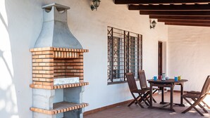 An outdoor patio with a brick oven, a table with chairs, and a decorative window grille. - Air-conditioned Holiday Home with Patio, Pool, Wi-Fi and Lawn (Barbate)