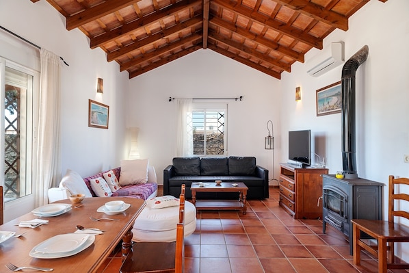 A cozy living room with a wooden ceiling, a sofa, a dining table set, a fireplace, and a TV.
