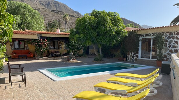 Piscina con sillones reclinables, zona de patio con sofá y mesa, y montañas al fondo.