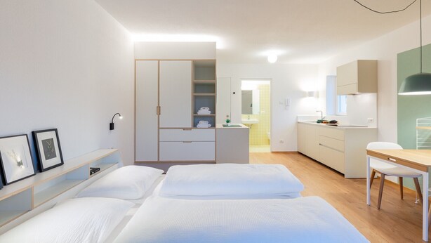 A hotel room with two beds, a desk, a kitchenette, and a bathroom visible through an open door.