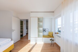 Un dormitorio con cama, ventana con cortinas, un banco empotrado con cojines y una pequeña mesa.