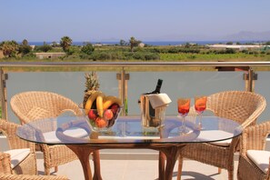 A glass-top table with wicker chairs, a fruit bowl, a bottle, and two glasses on a balcony overlooking a body of water and distant land.