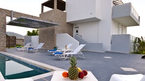 An outdoor pool area with lounge chairs, a table with fruit, and a modern building with stone and white walls. - Xenos Villa 1 With Private Swimming Pool Near The Sea (Kos)