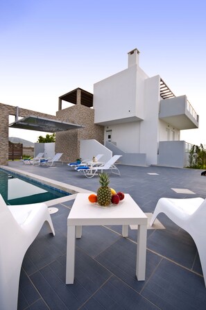 An outdoor pool area with lounge chairs, a table with fruit, and a modern building with stone and white walls.