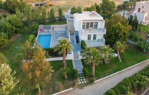 A modern two-story house with a swimming pool, surrounded by greenery and palm trees.