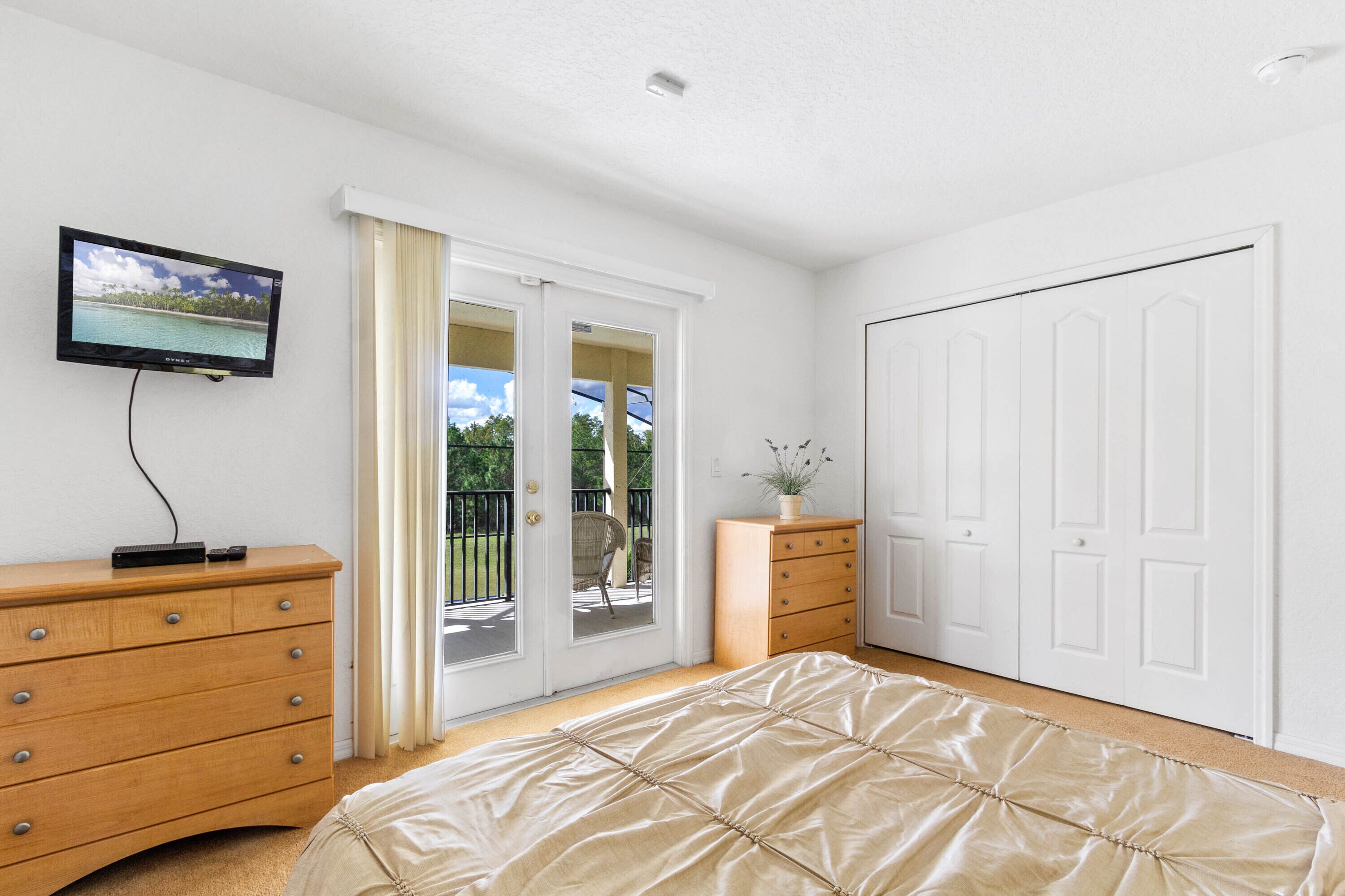 A bedroom with a bed, a TV, a dresser, a sliding glass door to a balcony, and a white wardrobe.