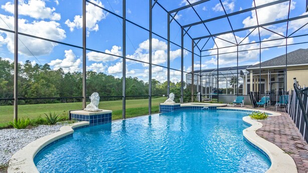Una piscina con dos estatuas, rodeada por una zona techada con sillas y vistas a una casa y árboles.