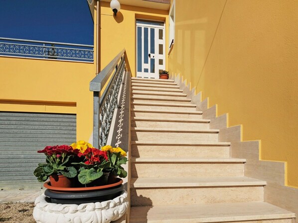 Una escalera conduce a una puerta con diseño azul y blanco, una planta en maceta con flores rojas y amarillas, y una pared amarilla.