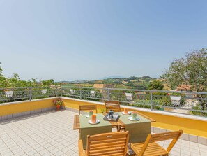 Una terraza con una mesa lista para dos personas, rodeada por un muro amarillo y una baranda metálica, con vistas a un paisaje pintoresco.