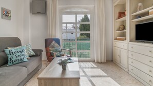 Un salon comprenant un canapé, un fauteuil, une table basse, un téléviseur et une grande fenêtre avec des rideaux.