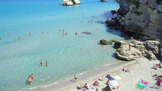 Una platja amb aigua cristal·lina, formacions rocoses i persones banyant-se i prenent el sol.