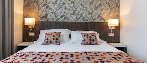 A hotel room with a large bed, two bedside lamps, a patterned wallpaper, and a geometric throw blanket.