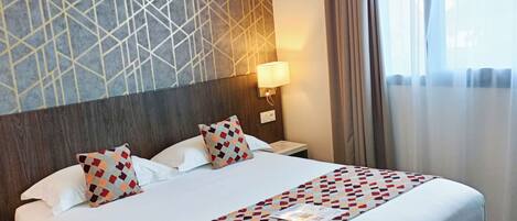 A hotel room with a large bed, a geometric-patterned wall, a nightstand with a lamp, and a window with curtains.