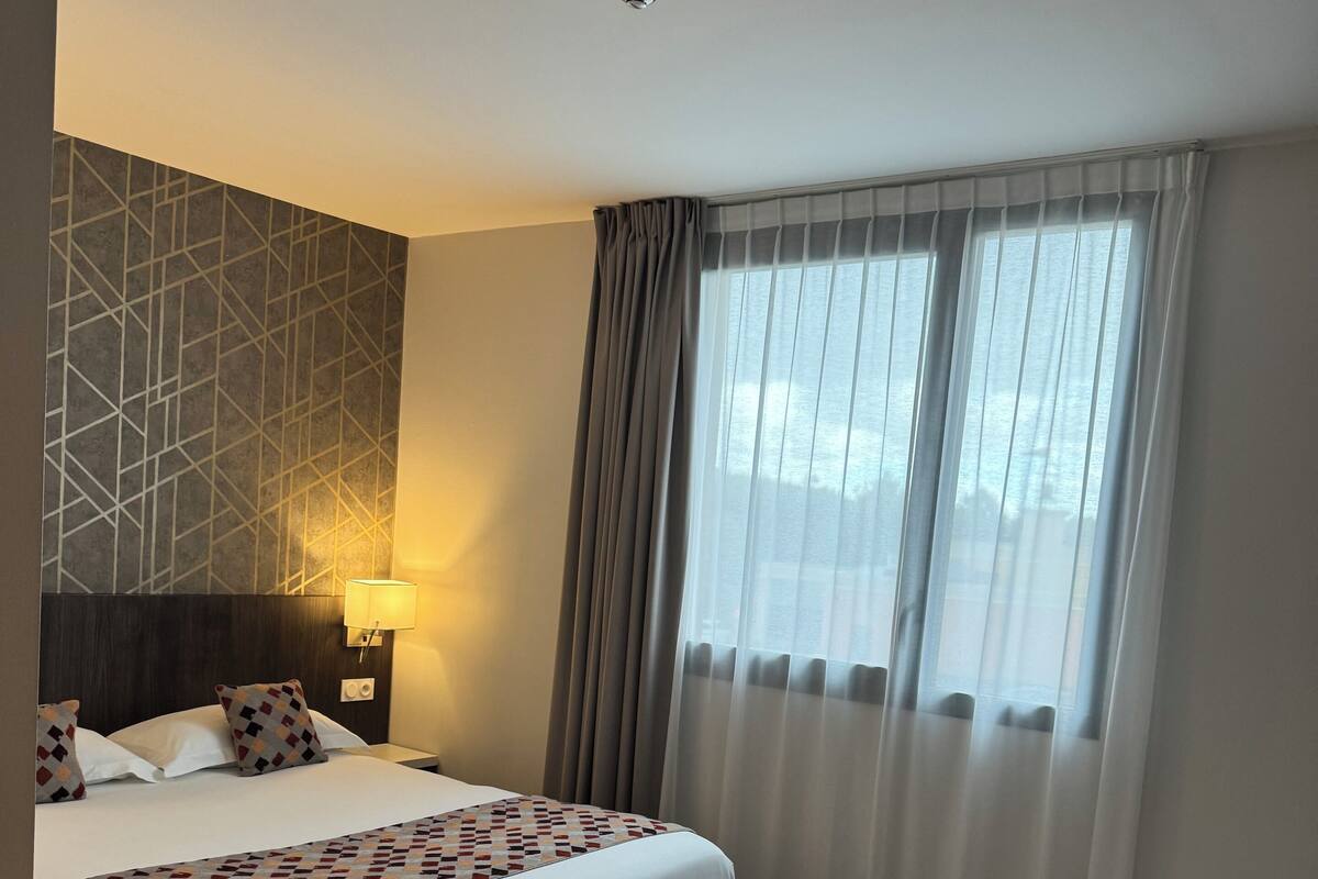Een hotelkamer met een groot bed, een kleiner bed, een nachtkastje met lamp, een wand met een geometrisch patroon en een raam met dunne gordijnen.