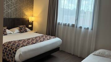 A hotel room with a large bed, a smaller bed, a nightstand with a lamp, a geometric-patterned wall, and a window with sheer curtains.