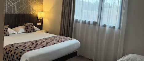 Camera d'albergo con un letto grande, un letto più piccolo, un comodino con una lampada, una parete con motivo geometrico e una finestra con tende trasparenti.