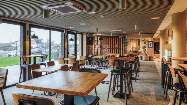 Un espace de restauration moderne avec des tables et des chaises en bois, de grandes fenêtres et une vue sur l’extérieur.