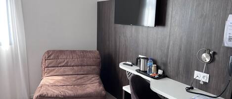 Une chambre d’hôtel moderne avec un fauteuil marron à coussins, un bureau doté d’un téléviseur à écran plat, une machine à café et une bouteille d’eau.