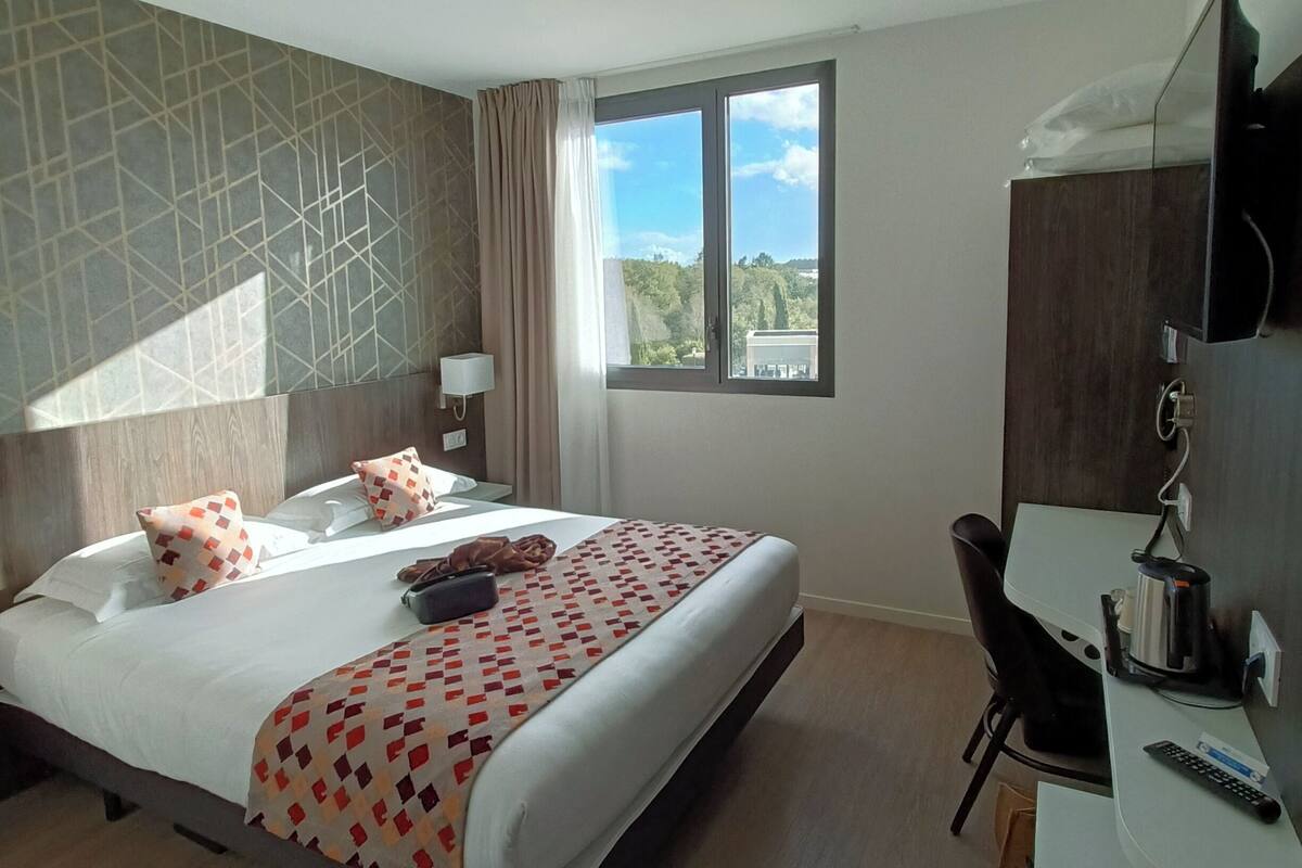 Een hotelkamer met een bed, een bureau, een stoel, een televisie en een raam met uitzicht op groen.