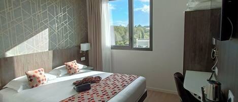 Habitación de hotel con cama, escritorio, silla, televisor y ventana con vistas a zonas verdes.