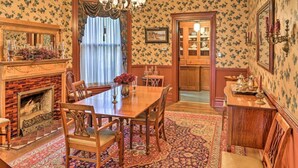 A traditional dining room with a brick fireplace, wooden dining table, and chairs. There is a patterned rug on the floor, and a chandelier hangs above the table.