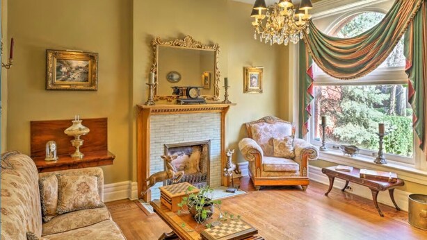 A traditional living room with a fireplace, a chandelier, and a view of greenery outside.
