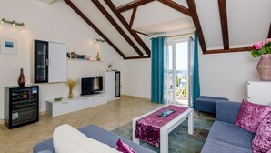 A modern living room with a high ceiling, built-in wooden beams, a flat-screen TV, a white cabinet, a glass-fronted refrigerator, a blue sofa, a purple cushion, and a view of the outdoors through a glass door. - Apartment Alba (Konavle)