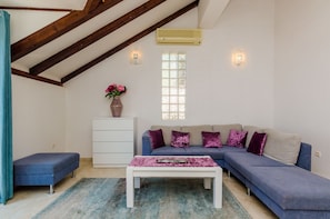 Un salon avec un canapé bleu, une table basse blanche, un banc bleu, un meuble blanc, une fenêtre à motifs géométriques et un appareil de climatisation.