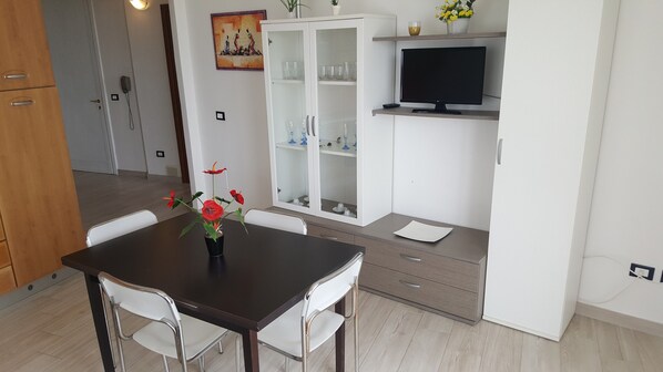 A modern kitchen with a dining table, chairs, a cabinet with glass doors, a television, and a potted plant.