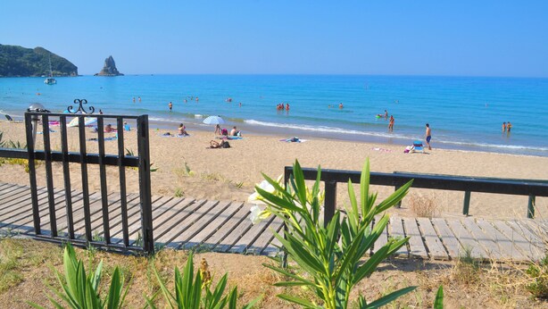 Ein Strand mit einer Holzpromenade, die zum Ufer führt, klarem, blauen Wasser und einer in der Ferne liegenden Felsformation.