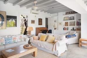 Een woonkamer met een witte bank, een houten salontafel en een lamp. Er hangen ingekaderde foto's aan de muur en er staat een boekenplank vol boeken.