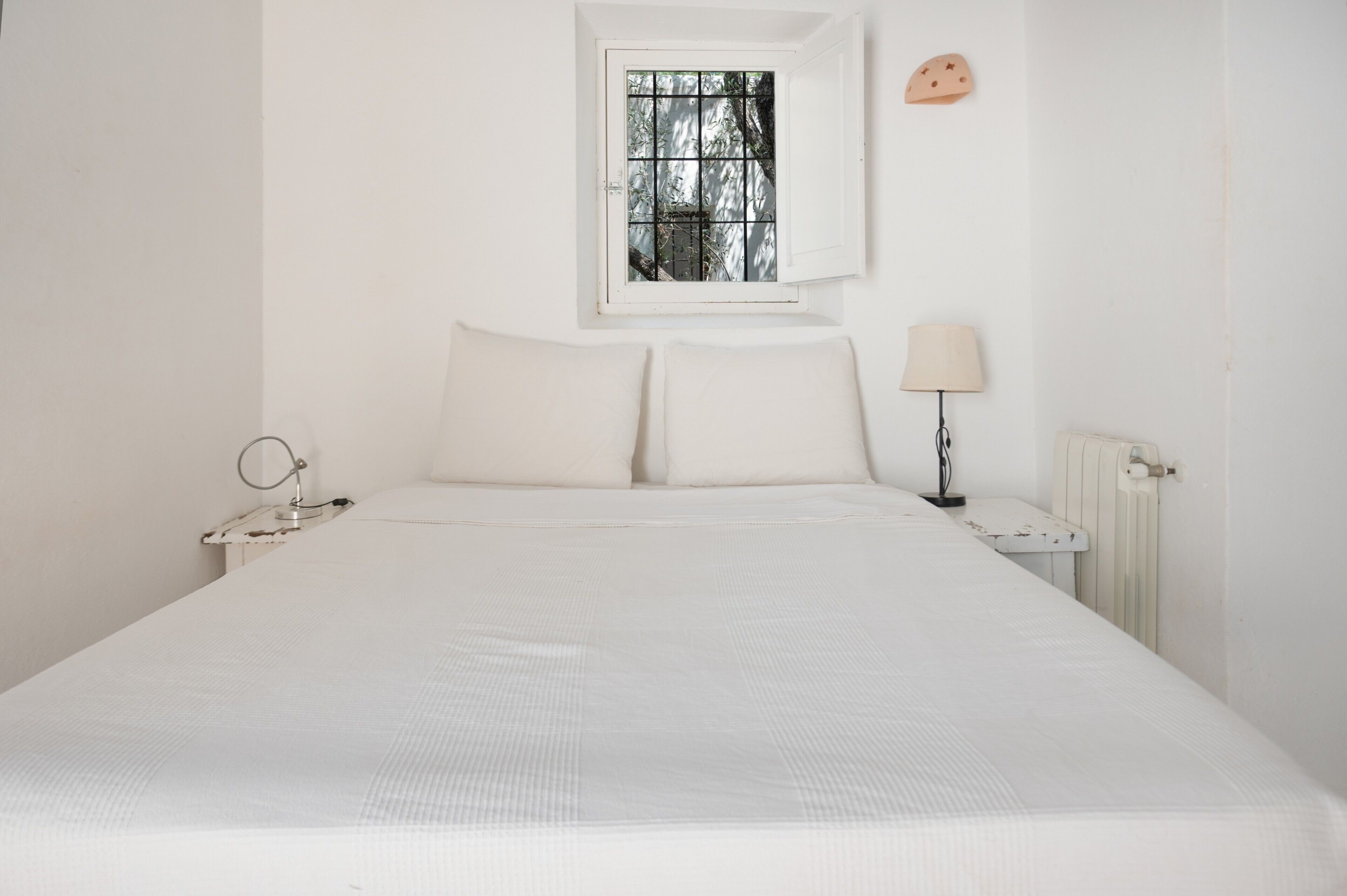 A white bedroom with a single bed, a bedside table with a lamp, a window with bars, and a wall-mounted decoration.