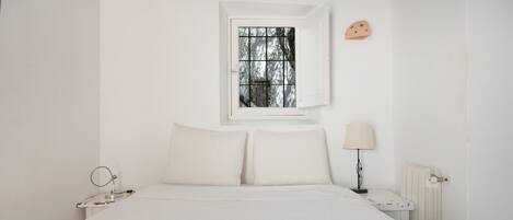 A white bedroom with a single bed, a bedside table with a lamp, a window with bars, and a wall-mounted decoration.