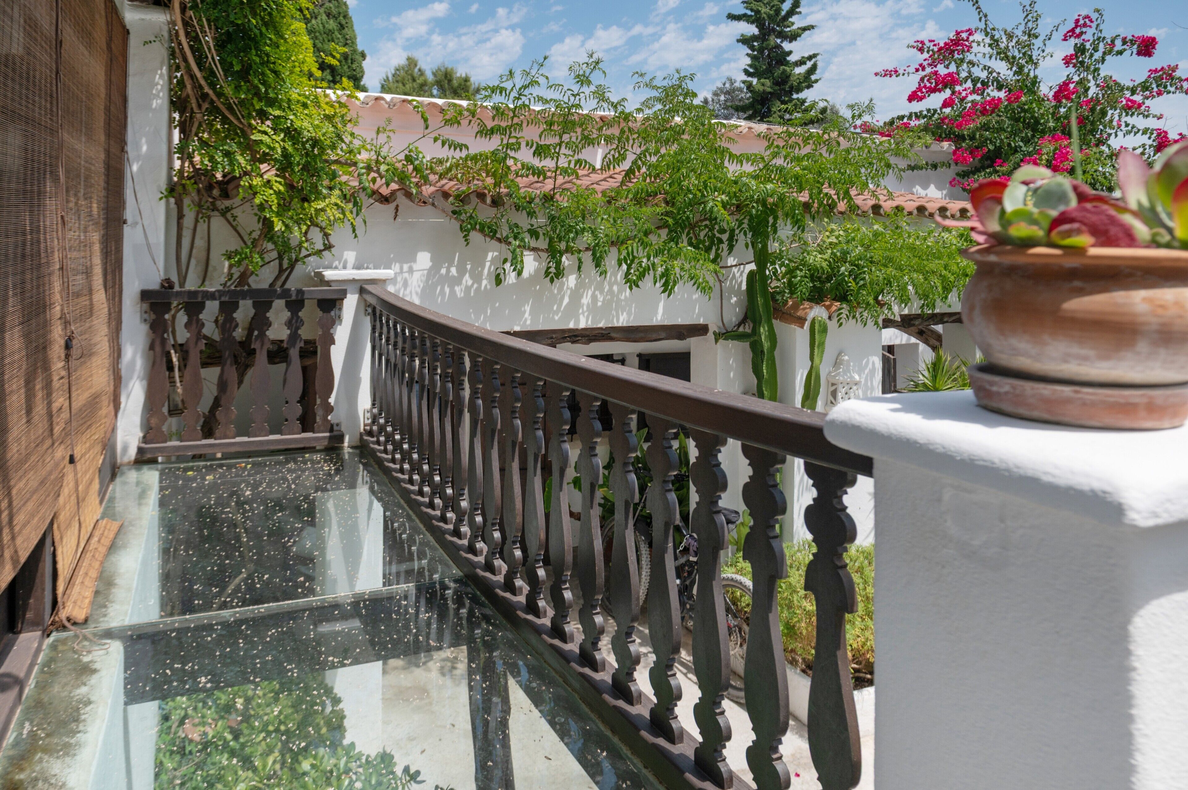 A glass balustrade with a metal railing, a potted plant, and a view of a building with a tiled roof and greenery.
