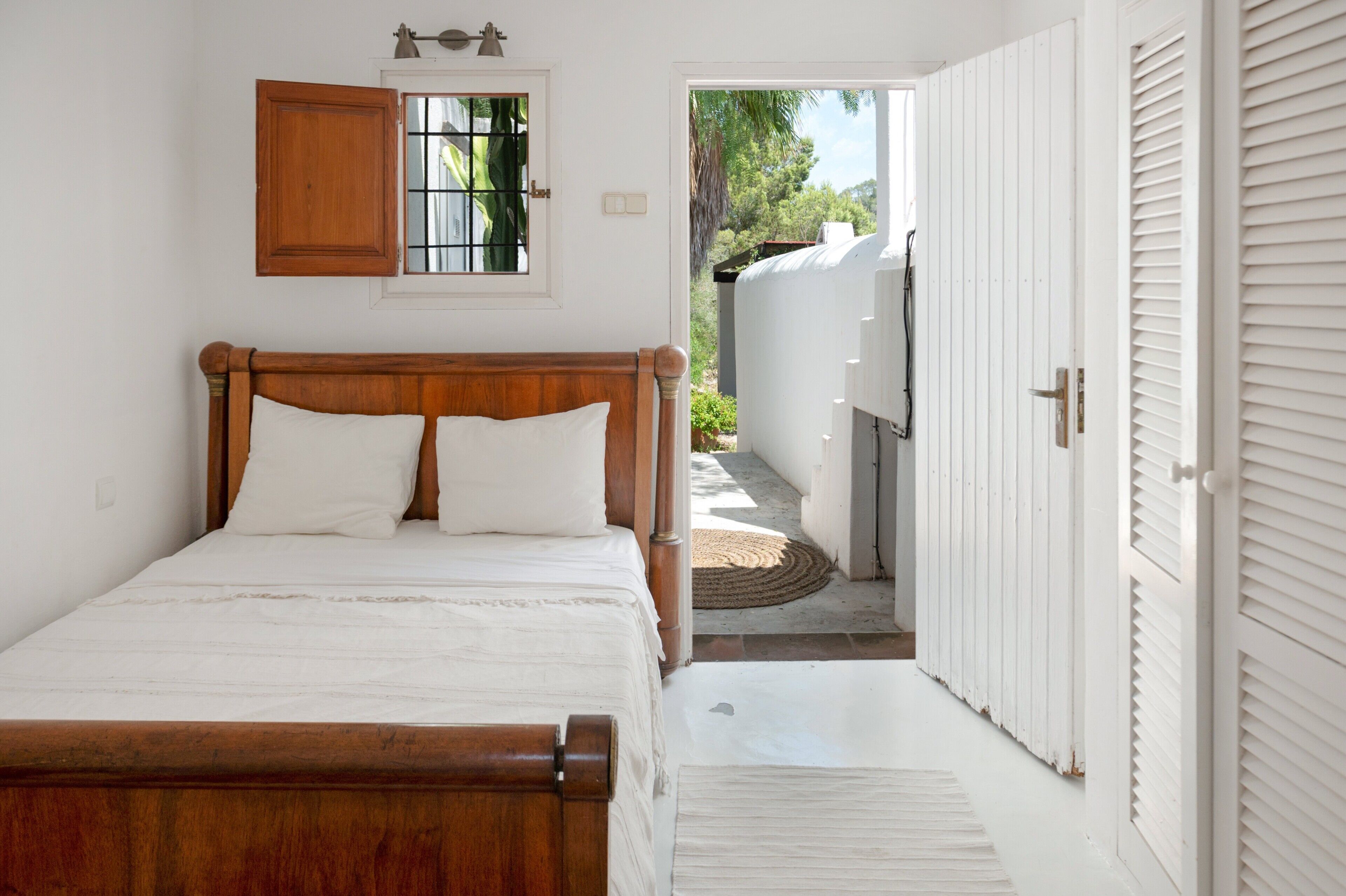 Un dormitorio con cama de madera, ventana con vista a follaje y puerta blanca que da al exterior.