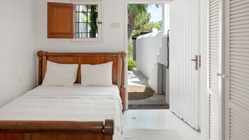 Un dormitorio con cama de madera, ventana con vista a follaje y puerta blanca que da al exterior.