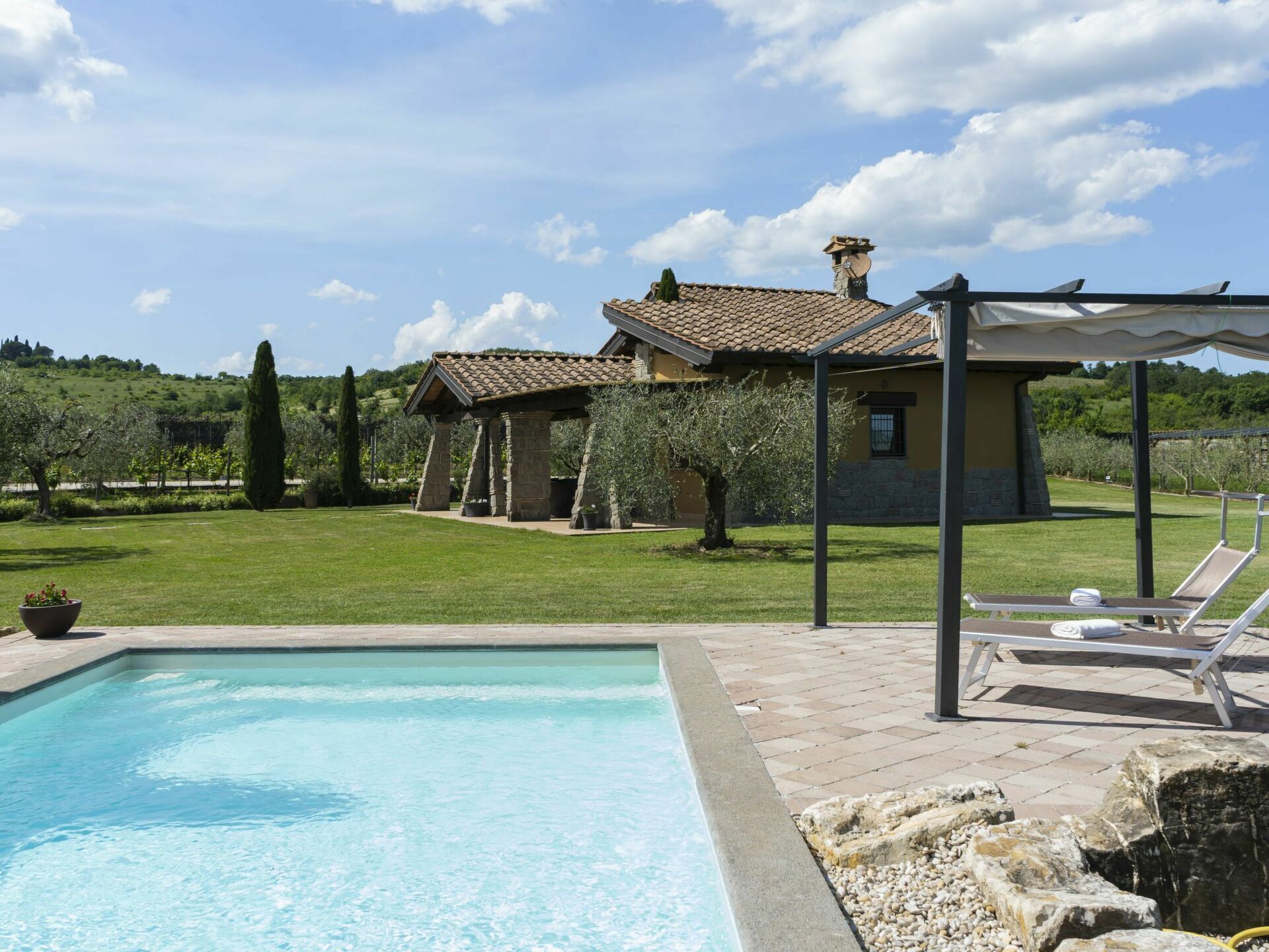 A swimming pool with a stone border, a grassy lawn, and a building with a tiled roof.