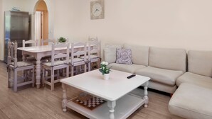 Een woonkamer met een witte tafel, bijpassende stoelen, een beige bank, een klok aan de muur en een kleine plant op de tafel.
