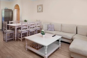 Un salon avec une table blanche, des chaises assorties, un canapé beige, une horloge au mur et une petite plante sur la table.