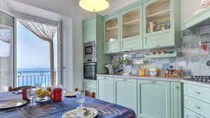 A kitchen with light green cabinets, a dining table set with plates, glasses, and fruits, and a view of the sea through a window. - Nice apartment for 6 people with WIFI, TV, terrace and pets allowed (Portoferraio)
