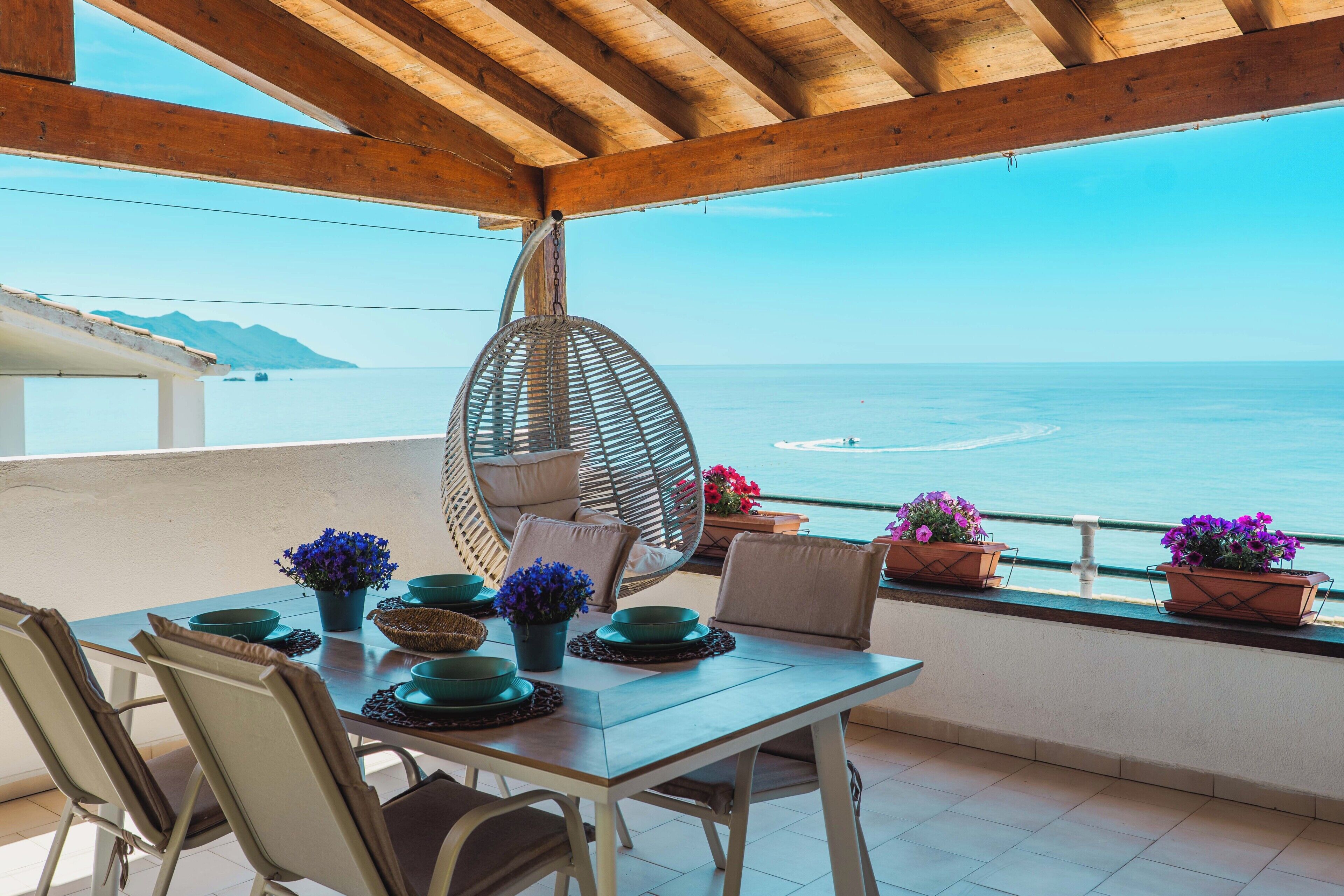A terrace with a wooden ceiling, a table set with green bowls and potted plants, and a view of the sea.