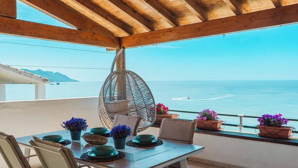 A terrace with a wooden ceiling, a table set with green bowls and potted plants, and a view of the sea.