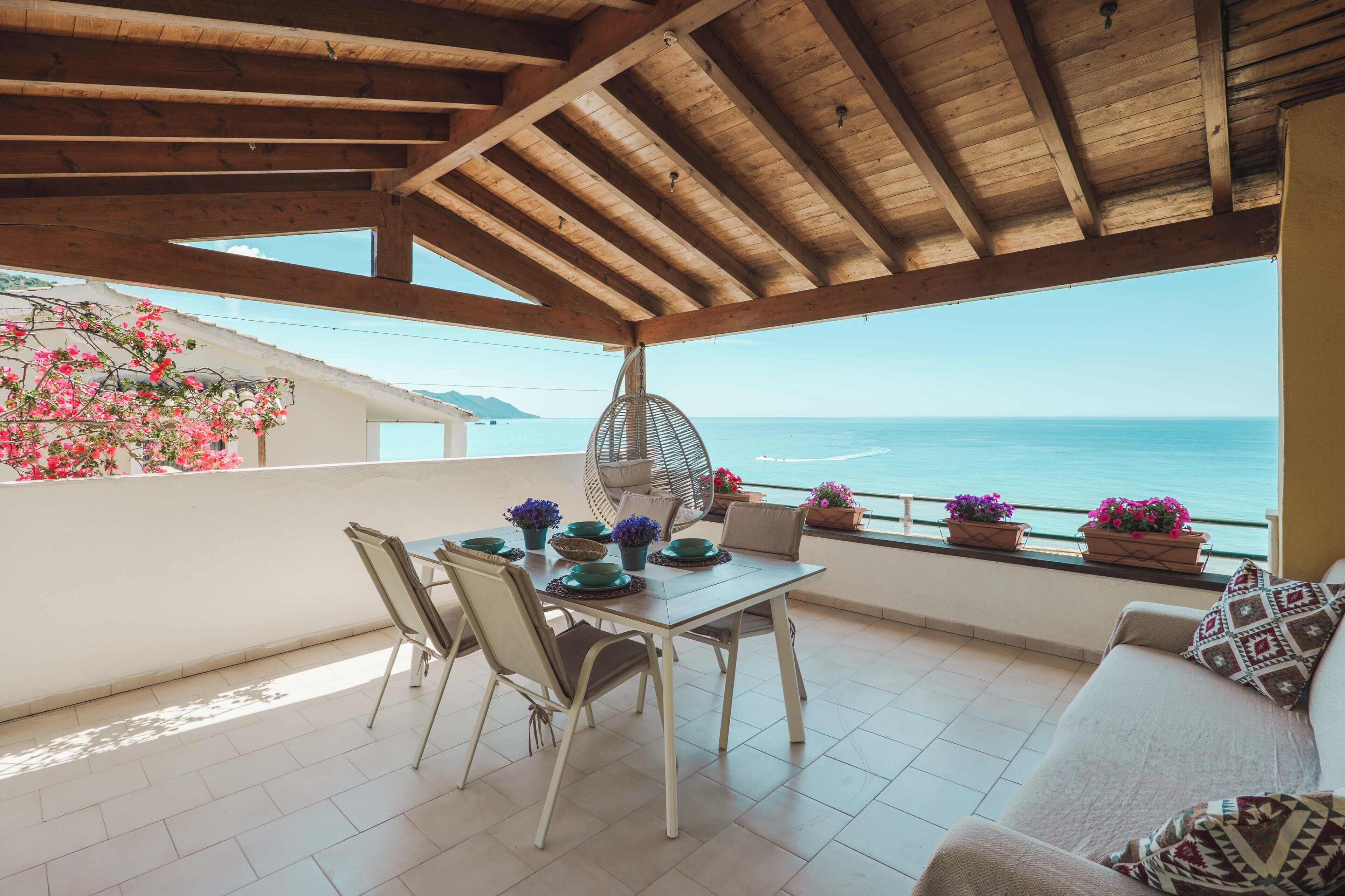 A covered outdoor seating area with a table, chairs, and a sofa, overlooking a body of water.