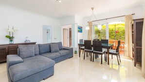 Una sala de estar moderna con un sofá seccional, una mesa de comedor, sillas de mimbre y vistas a un jardín a través de grandes ventanales.