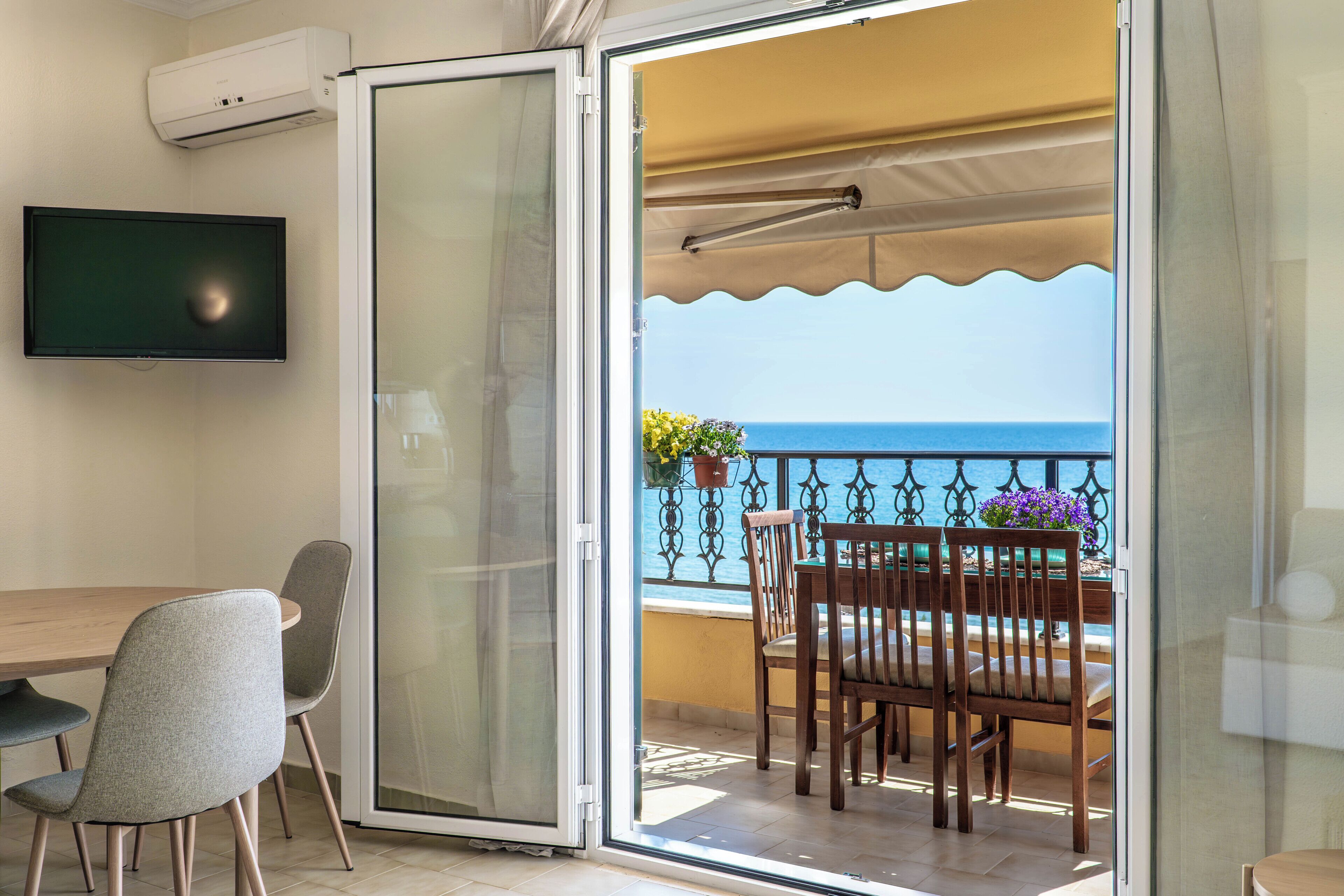 A balcony with a table and chairs, a TV mounted on the wall, and a view of the sea.