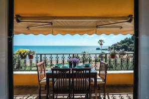 A balcony with a table and chairs, overlooking a coastal view with the sea and greenery.