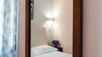 A hotel room with a large mirror, a bed with white linens, a nightstand with a lamp, and a window with curtains.