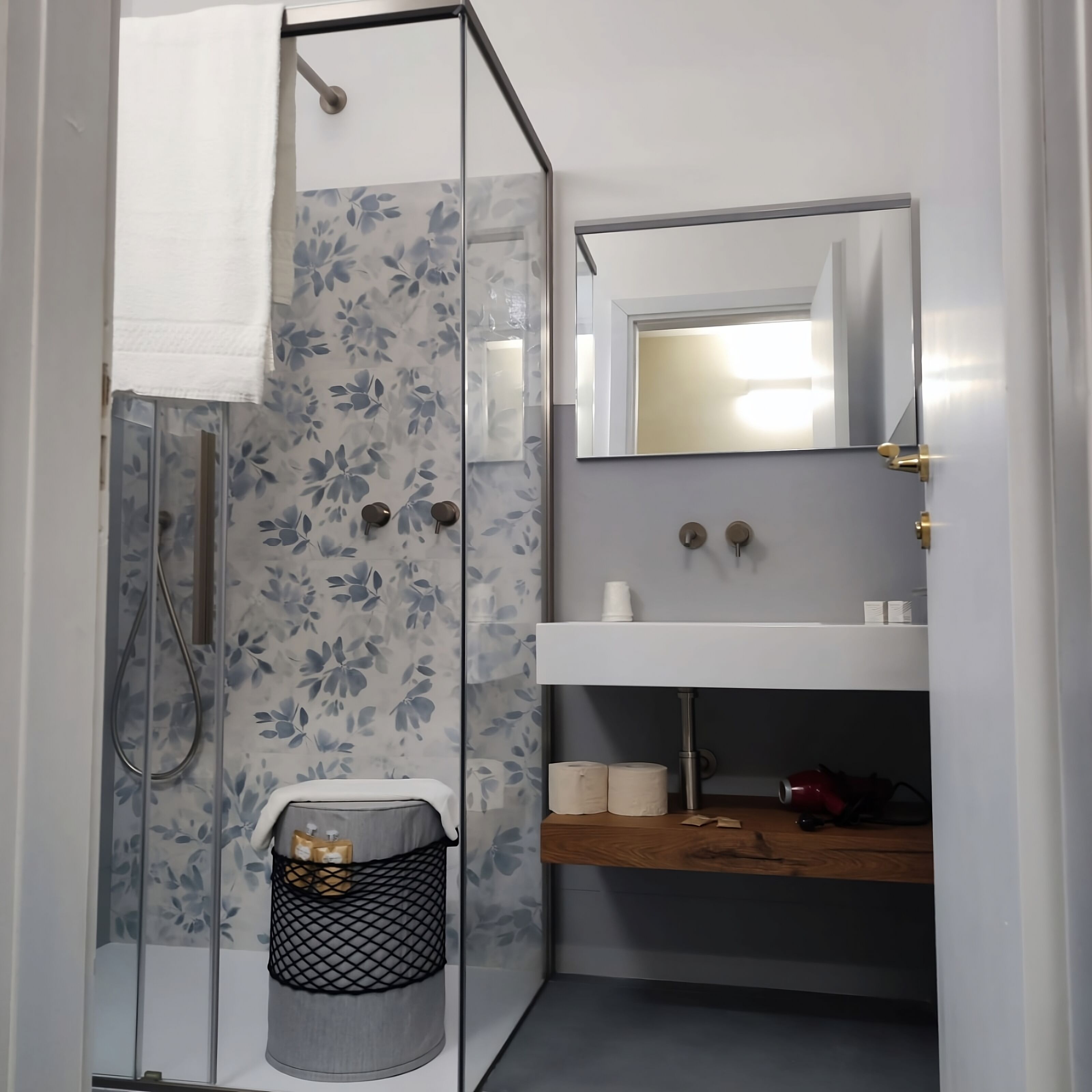 A bathroom with a glass shower enclosure, a white sink with a mirror above it, and a wooden shelf below.
