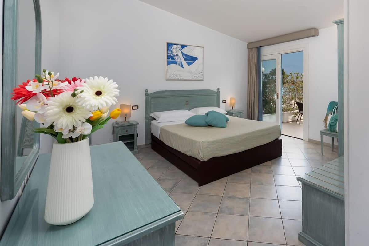 Un dormitorio con cama, mesita de noche, jarrón con flores, un cuadro en la pared y una puerta corrediza de vidrio que da a un espacio al aire libre.