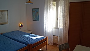 Een slaapkamer met een tweepersoonsbed, een stoel, een bureau, een raam met gordijnen, een wandlamp en een deur.